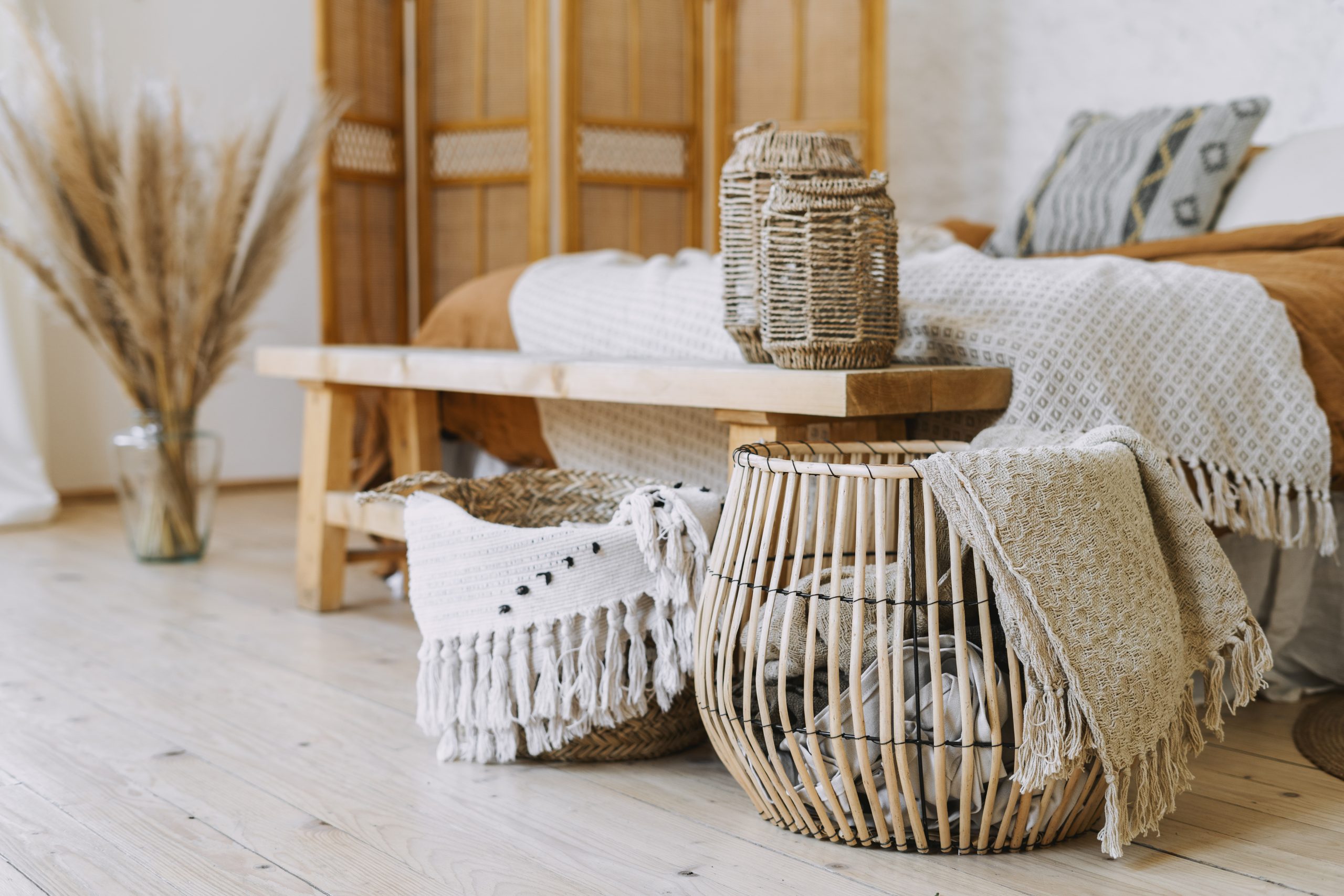 Selective focus on home decor. Comfortable bedroom in bohemian interior style with textile sheet on bed, wooden bench seat, bamboo dressing screen, dry plants in vase, wicker basket